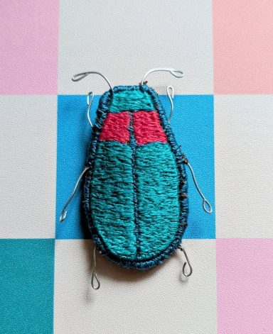 A handcrafted beetle figurine with blue and red detailing on a pastel background.